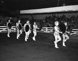 Drill team of women wearing shorts and holding round disks walk through the arena at the 1960 Little International Exposition at South Dakota State College. The audience watches from the stands.