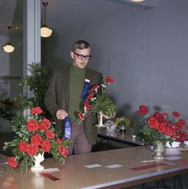 Man holding a floral arrangement at the 1971 Little International exposition at South Dakota State University. There are many arrangements on the tables around him.