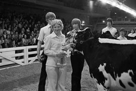 Women hold a trophy while standing by her dairy cow at the 1977 Little International Agricultural Exposition at South Dakota State University. Other people and cows stand behind her.