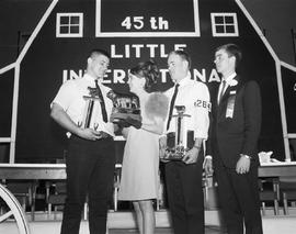 Little International Queen handing trophies to winners on the stage at the 1968 Little International exposition at South Dakota State University. The false barn wall is in the background.