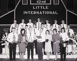 Group of winners hold their trophies and stand in front of the stage at the 1965 Little International Exposition at South Dakota State University. The false barn wall is in the background.