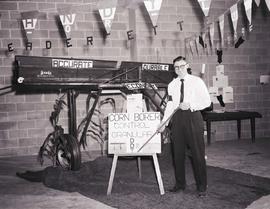 Men stands in front of farm machinery with signs that read, "Accurate, durable economy," at the 1956 Little International Exposition at South Dakota State College. He has a stick pointing at a sign that reads, "Corn borer control granulat DDT."