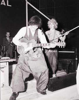 Two clowns on stage entertaining the audience at the 1964 Little International Agricultural Exposition at South Dakota State University. One clown is playing a guitar, the other is pretending to play a rake like a guitar.