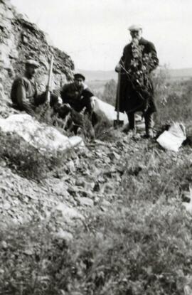 N.E. Hansen and two assistants climb rough terrain to gather specimens in their search for hardy peach trees in Mendoche in northern China in 1924, Hansen is holding a peach tree specimen, one man is holding a gun; written in pencil on the back: Mendoche, North China 1924.