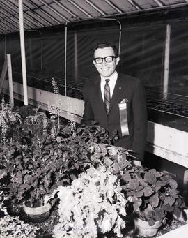Man with a ribbon on his jacket stands by potted plants in a greenhouse during the 1966 Little International exposition at South Dakota State University.