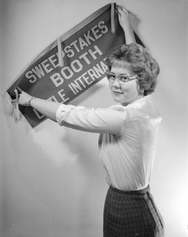 Queen, Lorraine Haverly, of the 1962 Little International Exposition at South Dakota State College holds a banner against the wall that reads: Sweepstake Booth Little International