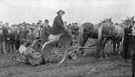 N.E. Hansen (wearing black collared overcoat and a hat) is standing by two horses drawing an unknown type of machinery, possible a planter of some king, there are three men, one driving and two sitting on the planter, the photograph was taken while N.E. Hansen was among the Kirghiz Tartars in Semipalatinsk, Siberia.