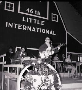 Man plays guitar and sings into the microphone on the stage the 1969 Little International exposition at South Dakota. Behind his are staff sitting at tables and the false barn wall.