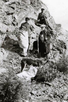 N.E. Hansen and two assistants gather specimens in their search for hardy peach trees in Mendoche in northern China in 1924, Hansen is holding a peach tree specimen, one man is holding a gun, burlap bags are holding specimens, there is a dog in the foreground; written in pencil on the back: Mendoche, North China 1924.