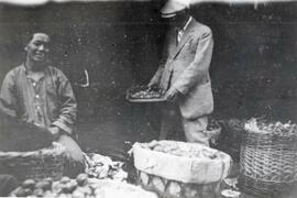 Man smiles for the camera while N.E. Hansen is looking the fruit he has for sale at a bazaar in Harbin, China; written in pencil on the back: Harbin 1924.