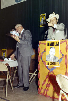 Frank Denholm is speaking at a campaign event. A man standing beside his is holding a cake.