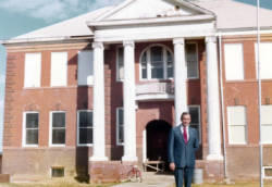 Frank Denholm standing in front of an abandoned school house. There is a bicycle on the sidewalk in front of the building.