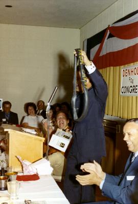 Frank Denholm at a podium during a campaign event. He is holding what looks like a flat bicycle intertube with writing on it.