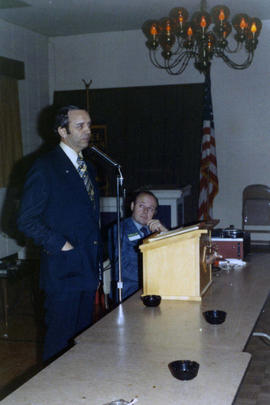 Frank Denholm standing at a podium an speaking to a group. There is a man seated to his left.