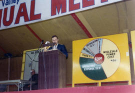 Man speaking at a podium on a stage. There is a large banner haning above him that appears to say Sioux Valley and Annual Meeting.