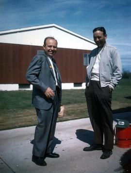 Frank Denholm (right) with a standing outdoors. There is a brown and white building in the background.