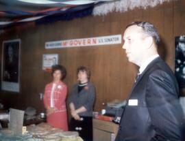 Frank Denholm at a campaign event in Aberdeen, South Dakota. Two women are standing by a table with pies on it. There is a McGovern U.S. Senator hanging on the wall.