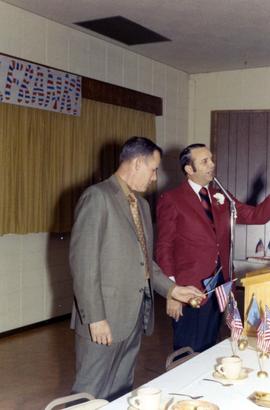 Frank Denholm speaking at a podium during his birthday party. A man is standing to the left.