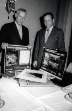 Frank Denholm and a man are in Denholm's office. They are holding posters about automated vegetation mapping and computer identification of terrain features.