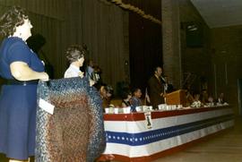 Frank Denholm speaking at a campaign event in an auditorium. Richard Kneip is seated to Denholm's right. There is a man with an accordian sitting on the stage behind Denholm.