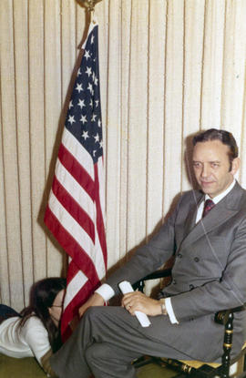 Frank Denholm seated in a chair next to the American flag. There is a woman in the floor holding on the the flag staff.