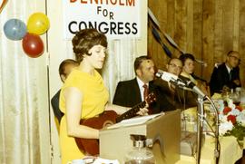 Woman playing a guitar at a podium during a campaign event for Frank Denholm.
