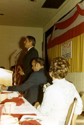 Man at podium speaking at a campaign event. Frank and Millie Denholm are seated to his left.