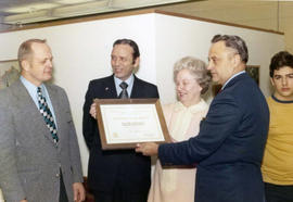 Frank Denholm receives a Certificate of Merit plaque from a group of people.