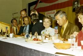 Frank Denholm talking with men surrounding him at a table during a campaign event. Millie Denholm is smiling.