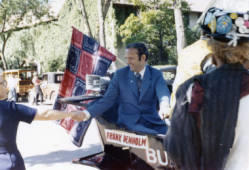 Frank Denholm shaking hands with a constituent while riding in the South Dakota State University Bummobile during the Hobo Day parade the hobo flag of peace is behind him