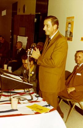 Richard Kneip at a podium speaking to a group at a campaign event. Frank Denholm ia seated behind him.