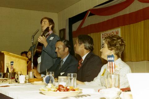 Man playing a guitar at a podium during a campaign event for Frank Denholm. Frank is seated to his left Millie Denholm is seated on the far left.
