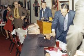 Frank Denholm shaking with a man at an event. A man is standing at the podium in the background.