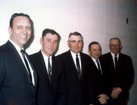 Frank Denholm with four men. They are standing in front of a white concrete block wall.