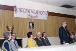 Man speaking at a podium during a campaign event for Frank Denholm. Denholm is seated behind him. There is a banner hanging on the wall which says Congratulations Frank.