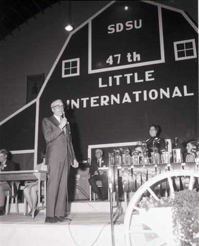 Man holding a microphone speaking to the audience on the stage at the 1970 Little International exposition at South Dakota State University. The trophy table and the false barn wall are in the background.