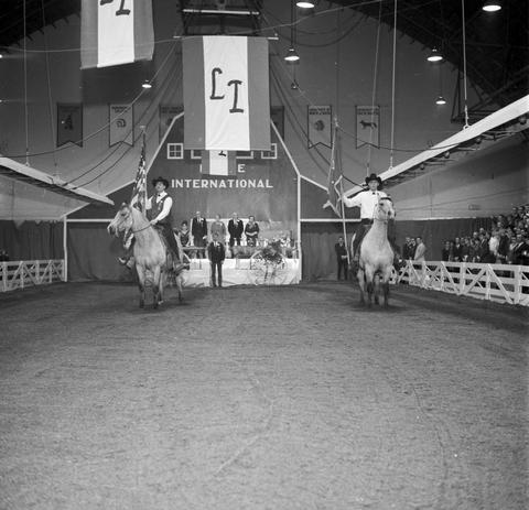 Two men on horseback serve as the color guard presenting the U.S. and State of South Dakota flags at the 1967 Little International exposition at South Dakota State University. The stage and false barn wall are in the background.