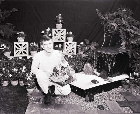 Man seated at the horticulture booth at the 1964 Little International Agricultural Exposition at South Dakota State University. He is surrounded by house plants.