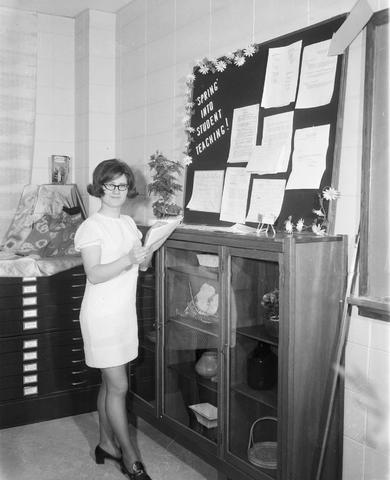 Woman stands by a display case at the 1970 Little International exposition at South Dakota State University. A bulletin board is above the case with words that read: Spring into student teaching!