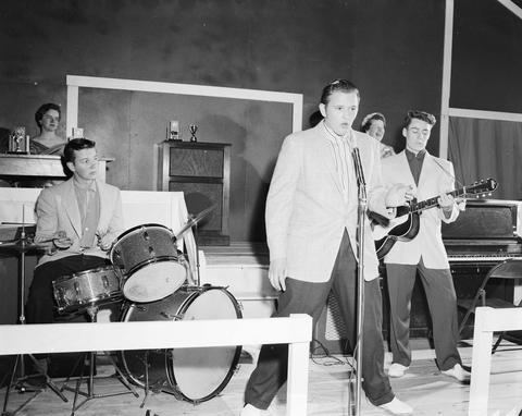 Three men entertain on the stage of the 1957 Little International Exposition at South Dakota State College. One man is singing, one is playing the guitar, and one is playing the drums.