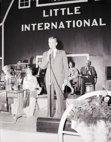 Announcer on stage speaking into a microphone at the 1964 Little International Agricultural Exposition at South Dakota State University. The Little I Queen is seated at a table behind him. There are trophies on the table.