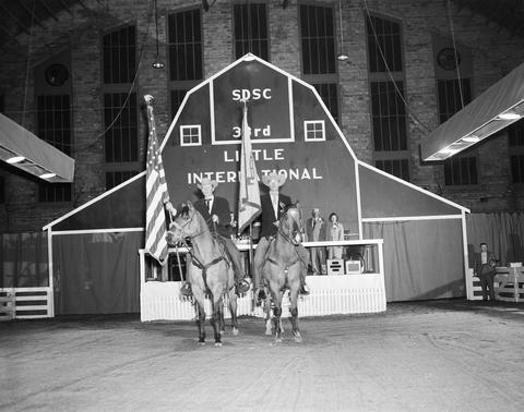 Two men on horseback holding a U.S. flag and a State of South Dakota in the arena at the 1956 Little International Exposition at South Dakota State College. The false barn wall is behind them.