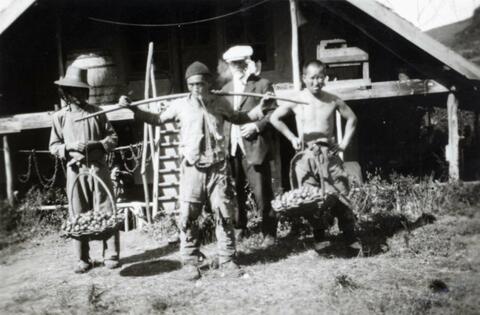 N.E. Hansen is standing behind a man with a carrying pole on his shoulders and two baskets full of pears at Hansen's headquarters while conducting pear research at Saolin in northern China, there are two men accompanying them; written in pencil on the back: North China 1924.