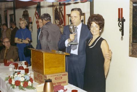 Frank Denholm standing behind a podium holding a microphone. His arm is around the woman standing next to him.