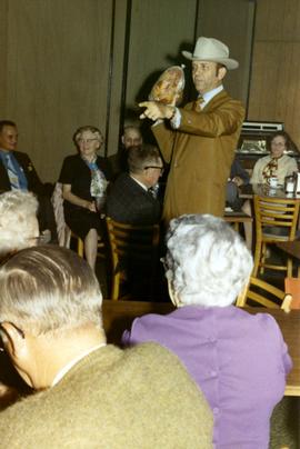 Frank Denholm at a campaign event. He is holding a ham while the audience is seated on chairs around him.