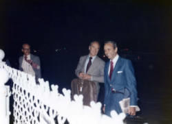 Frank Denholm and George McGovern standing by an airport fence at night. They are both holding luggage.