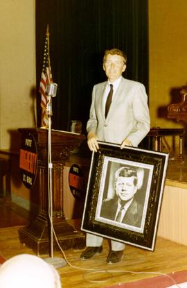 William Dougherty, Richard Kneip's running mate in the 1970 election, isstanding next to a podium holding a framed picture of John Kennedy at a campaign event. The podium has a sign attached to the front saying Vote Dougherty Lt. Governor.