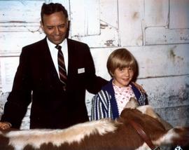 Frank Denholm is on the campaign trail. He is in a barn with child. They are standing next to cow.