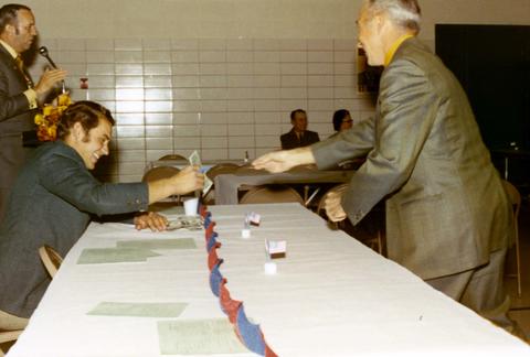 Frank Denholm speaking at a podium during a event. A man (standing) is handing another man (seated) a dollar bill in the foreground.