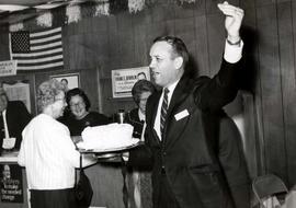 Frank Denholm at a campaign event in Aberdeen, South Dakota. He is holding a white frosted cake in his hand and appears to be auctioning the cake to the highest bidder.